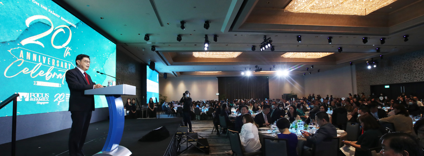 Man at podium for Focus on the Family Singapore's 20th Anniversary celebration. Audience seated at tables.